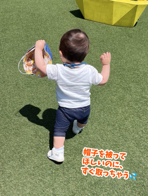 一歳一ヶ月頃の息子のりん太の写真。公園の芝生の上をバンザイで歩いています。被せてあげた帽子を取って片手に持っています。
